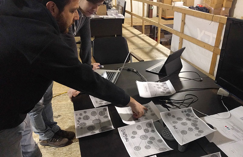 Two people map how information flows through a factory in a tabletop exercise: printed diagrams for each area, one marked Shipping, with a laptop, a tablet and a TV for supplemental data
