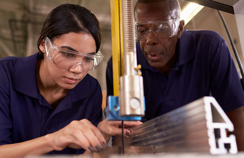 A trainer, mid-sentence, coaches a newer operator lining up an aluminum workpiece at a vertical band saw; both wear safety glasses.